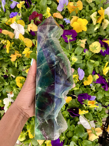 Large Rainbow Fluorite tower held over a flower garden. Fluorite is known to be a highly protective stone, especially on the psychic level. Fluorite helps you filter through negative energies.