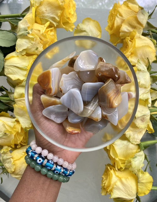 Banded Agate is associated with the Root Chakra and is a grounding stone. It helps to harmonize our bodies and souls so that we may focus on the present moment. Tumbled Banded Agate in a clear bowl is pictured .
