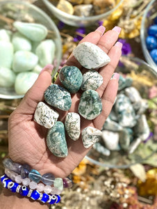 Tree Agate is a stone about abundance and grounding.  Tumbled Tree Agate pictured in palm of hand with bowls of crystals in the background.
